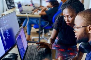 Black lady is helping a black gentleman with his laptop in a computer class.